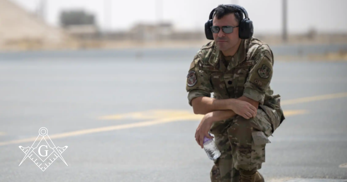 Lieutenant Colonel Brian Smith kneels on an airfield in desert conditions, wearing camouflage uniform, protective ear gear, and sunglasses, holding a water bottle. A Masonic square and compasses emblem is displayed in the corner.